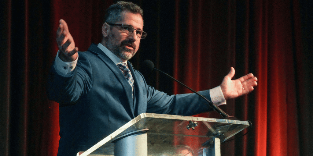 Wayne Whitzell speaking with open arms at a podium during a facility management event.